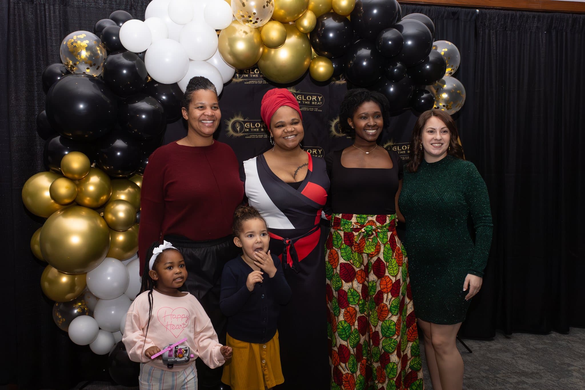 Four women and two girls pose by a black, white and gold balloon arch at the SneakerBall.